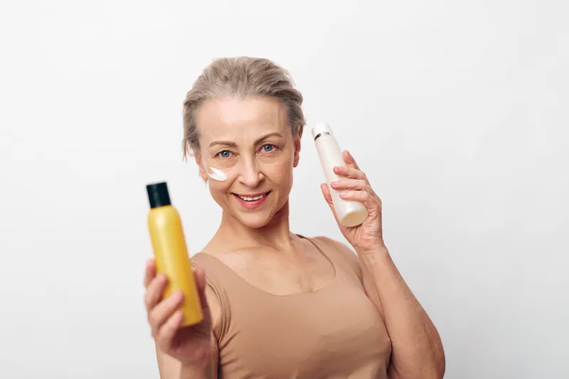 Smiling mature woman with skincare cream on her face holds two cosmetic bottles in hands against white background. Concept of beauty routine, anti aging, wellness and self care.