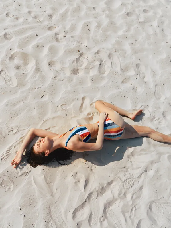Woman lies on sunlit sand wearing a striped bikini, relaxed pose with arms overhead, soft shadows and warm breeze creating a serene beach scene of summer calm.
