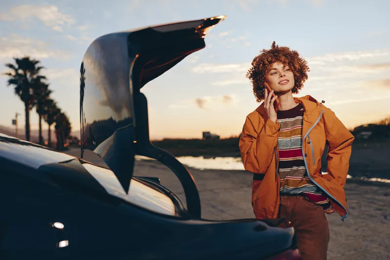 Woman lifestyle outdoors by a car at sunset, orange jacket and denim, curly hair smiling near open door, fashion travel for road trip adventure in warm light with evening backpack