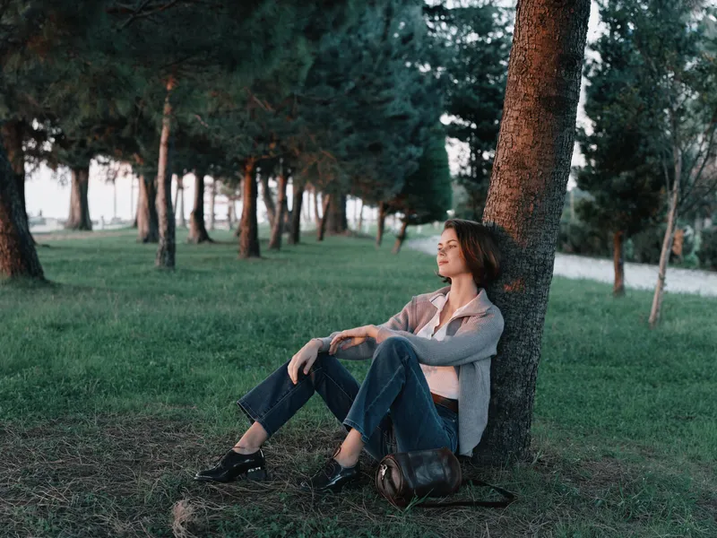 A relaxed woman sits against a tree in a peaceful park, enjoying quiet daylight. She gazes into the distance with a soft smile, creating a calm and reflective outdoor atmosphere.