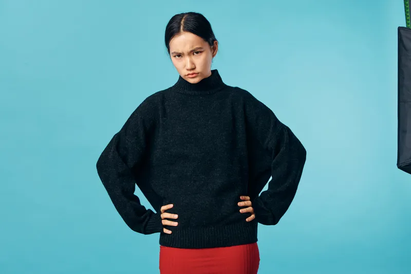 Young woman in black sweater with hands on hips, looking serious and focused against blue background. Female model wearing red skirt and dark outfit with confident posture.