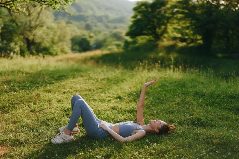 Young woman in activewear lying on grass in nature, expressing joy and relaxation in a vibrant outdoor setting during summer