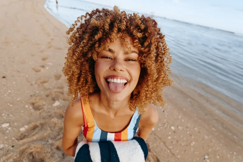 Young woman with curly hair laughs on a sunlit beach, looking toward the camera, wrapped in a striped towel, radiating carefree energy in a bright outdoor moment.
