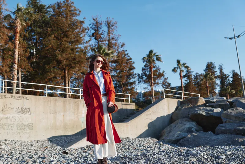 Young woman in a red trench coat walks confidently on a rocky beach under clear blue sky with sunglasses and casual white pants.