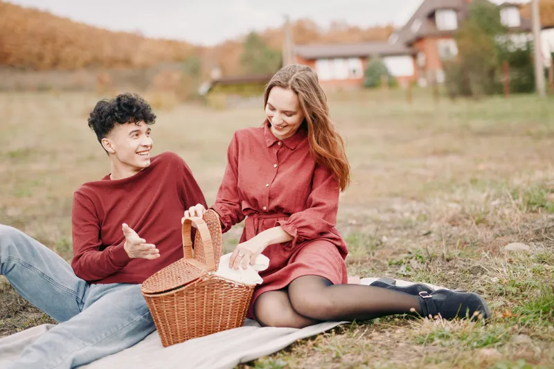 A candid outdoor picnic scene showing a young couple sharing bread from a wicker basket, genuine smiles and relaxed posture conveying authenticity and visual credibility in a rustic setting.