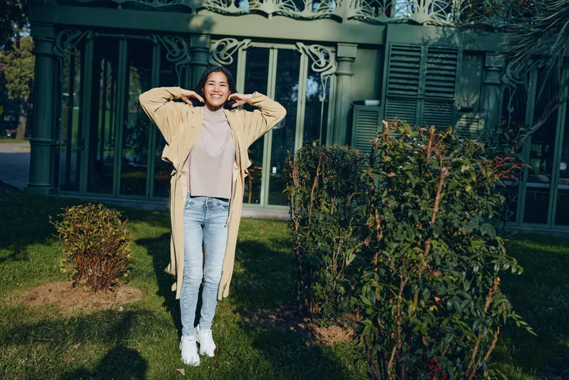 Young woman outdoors posing with hands behind head, wearing a long cardigan, jeans and sneakers in a sunny garden beside a house, radiating casual fashion and relaxed, joyful mood