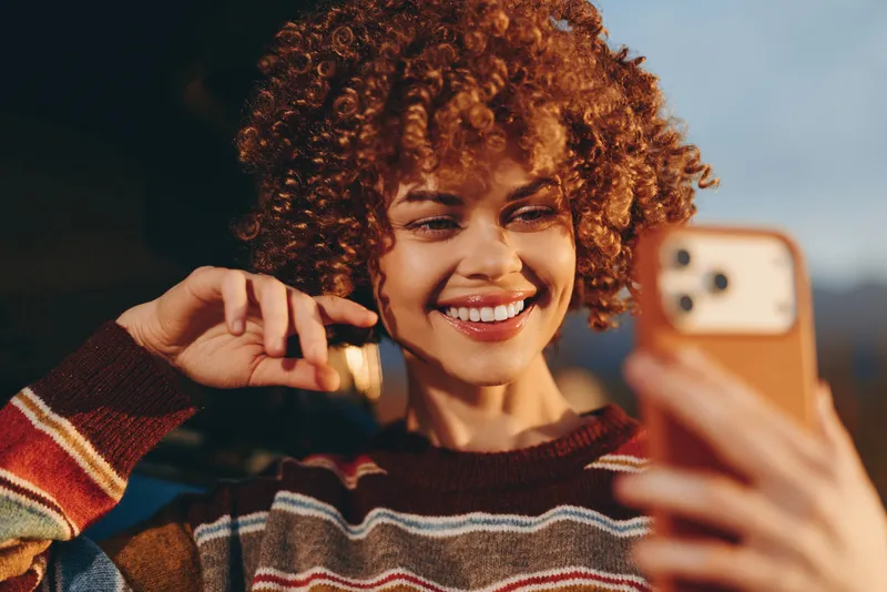 Smiling woman with curly hair making a selfie with a smartphone, wearing a cozy striped sweater. Outdoor lifestyle moment, close portrait with warm light and relaxed, happy mood.