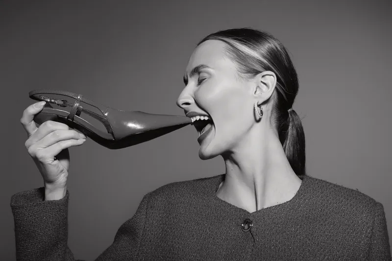 Smiling white woman with light skin holds high heel shoe near face in black and white studio photo. Concept of fashion, joy, and playful female model portrait.