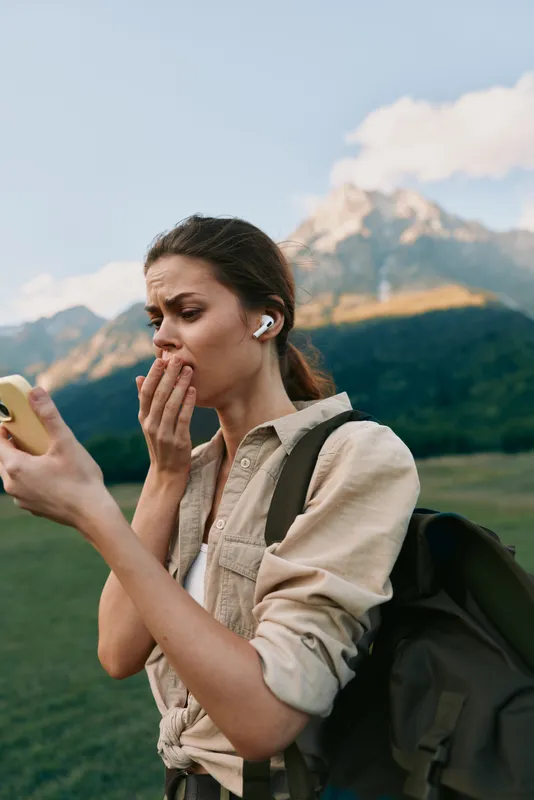 A young woman with a backpack looks shocked while checking her smartphone outdoors with mountains in the background during a clear day.