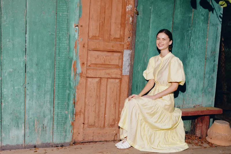 A woman in a yellow dress sits on a wooden bench beside a rustic teal door, smiling softly under natural light for a calm outdoor portrait.