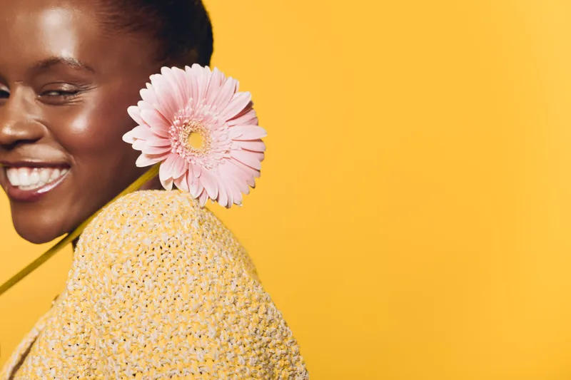 Happy African woman with dark skin smiles holding pink flower behind shoulder on yellow background. Concept of joy, natural beauty, floral, and positive mood in studio lighting.