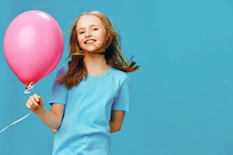 Happy girl with long hair holding a pink balloon in a blue background, wearing a light blue t shirt, expressing joy and playfulness in a vibrant setting