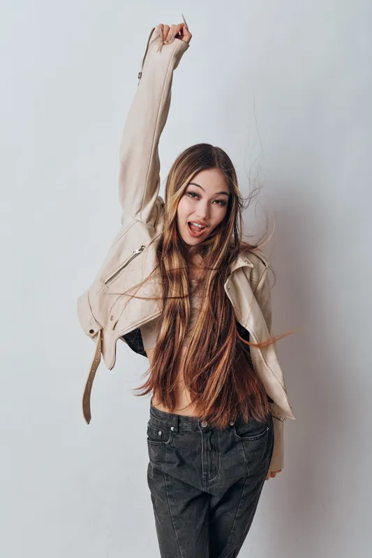 Young woman with long hair wearing casual jacket and jeans jumping energetically posing against white background with happy expression in studio portrait photo.