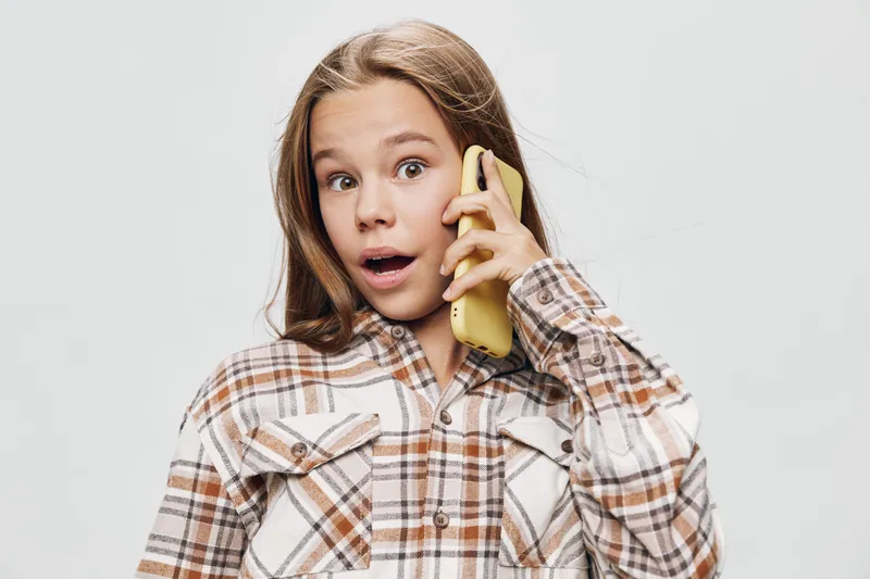 Girl with surprised expression talking on smartphone wearing checked shirt against white background. Young female with mobile phone in hand showing amazement or shock for communication.