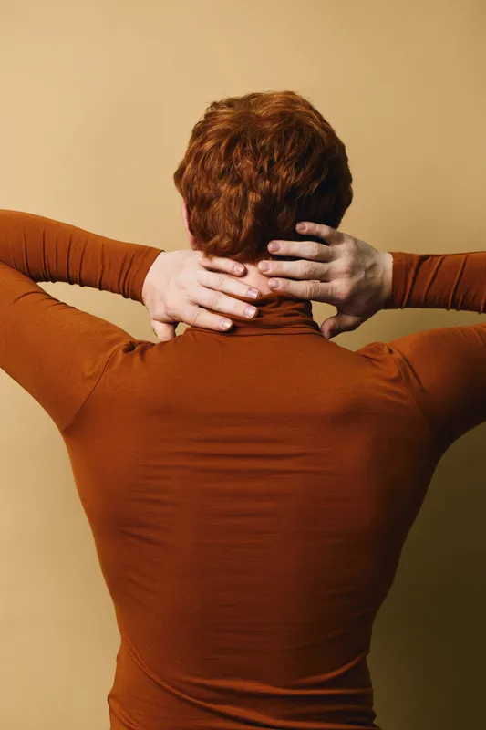 Person with red hair and brown long sleeve shirt standing back to camera with hands on neck against beige background for relaxation and stress relief concept.
