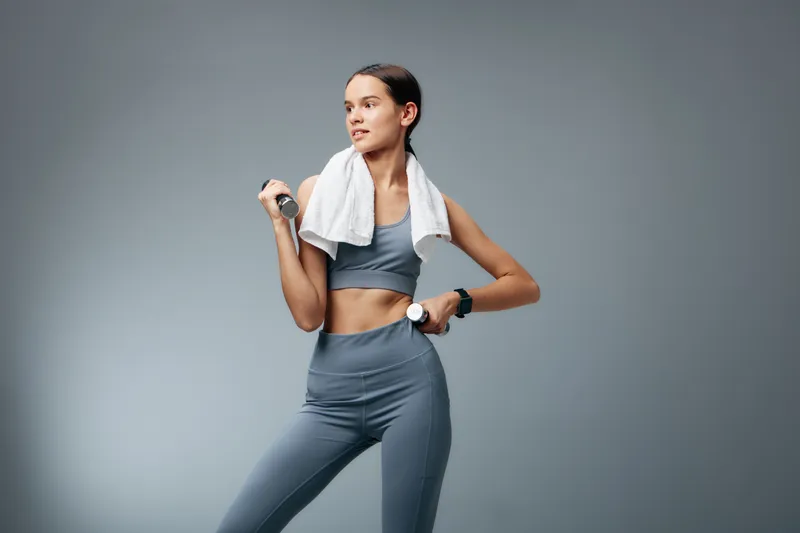 A athletic woman in grey workout wear holds a dumbbell with determination, a towel draped over her shoulders, posed against a clean studio backdrop for a dynamic fitness moment.