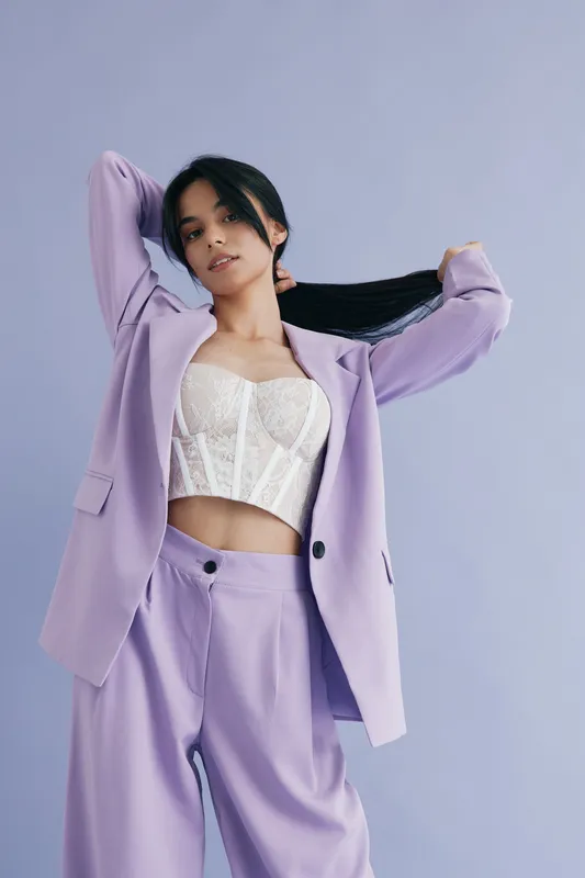 Young woman in lavender suit adjusting her hair, wearing a white lace corset top, standing against a soft purple background with a confident and stylish pose.