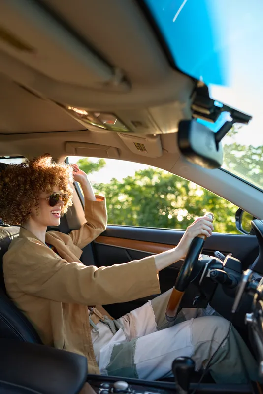 Woman driver seated in a stylish car, wearing sunglasses and a beige jacket, enjoying a sunny road trip, travel friendly casual lifestyle with retro film color vibe.