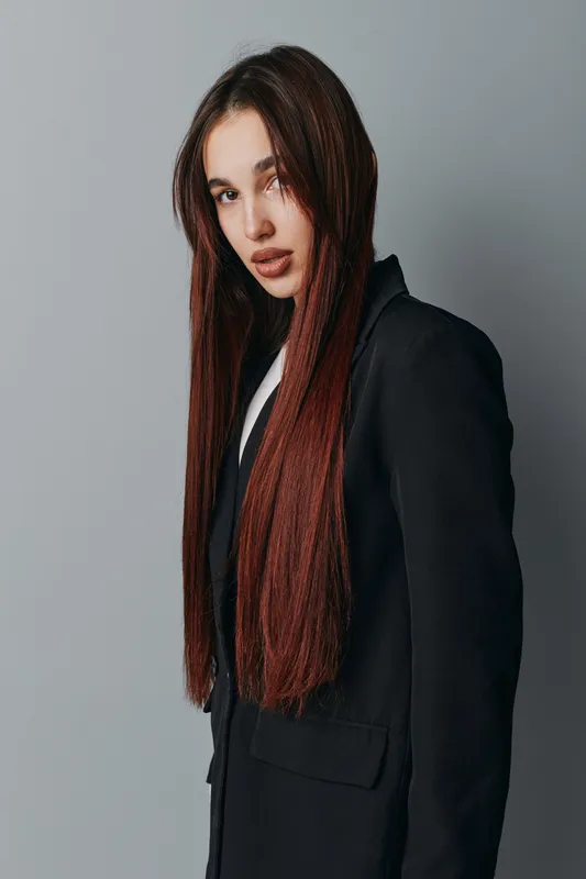 Young woman with long straight hair wearing black blazer standing against gray background looking at camera with confident expression in studio portrait. Female model with natural makeup and casual