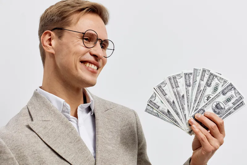 Smiling young caucasian man dressed in a beige suit and glasses holding a fan of hundred-dollar bills in his hand, looking happy and confident, isolated on plain white background. Business success con
