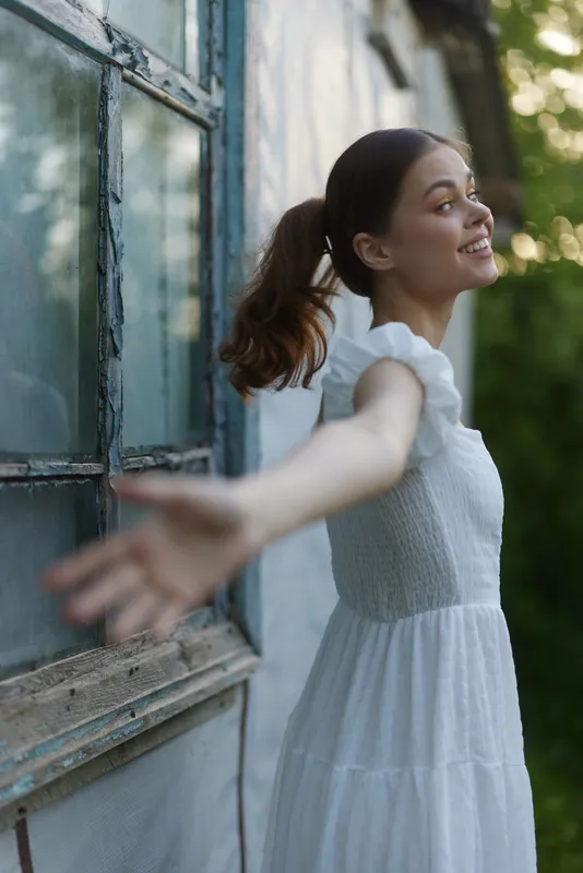 Young woman in a white dress joyfully embraces nature, expressing freedom and happiness near a rustic window in a lush green environment