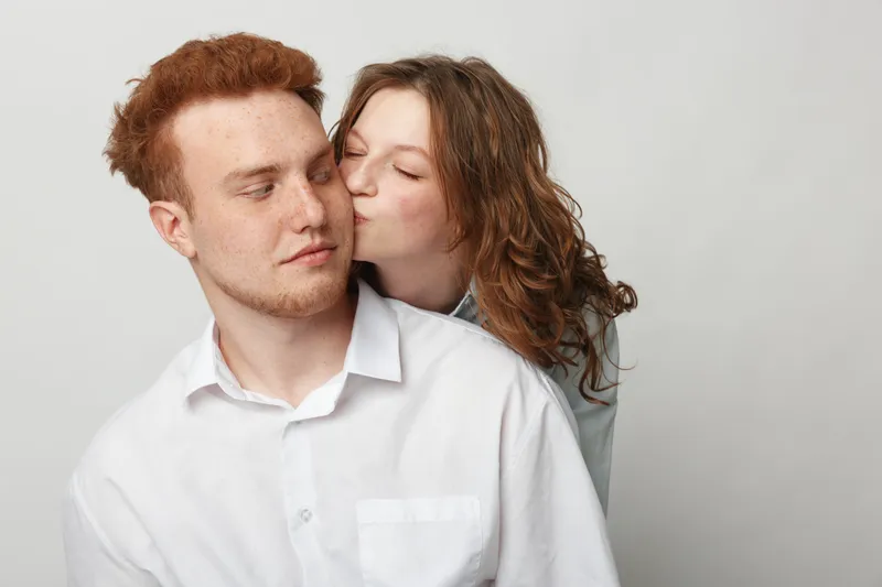 Young woman with curly hair kisses redhead man on cheek. Both wear white shirts against plain background. Concept of affection, love, intimacy, and relationship between couple.