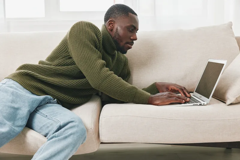 Young African man in a cozy green sweater working on a laptop at home, showcasing a focused expression in a bright and modern living room