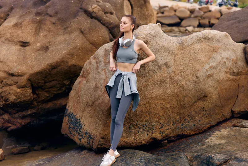 Fit young woman in activewear enjoying nature outdoors, standing on rocks near the beach, wearing headphones and a relaxed expression, perfect for healthy lifestyle themes