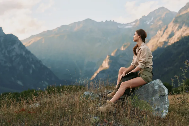 A woman in casual clothes sits on a rock in a mountain meadow, looking at the distant peaks during a calm sunset. Concept for lifestyle, nature, and relaxation.