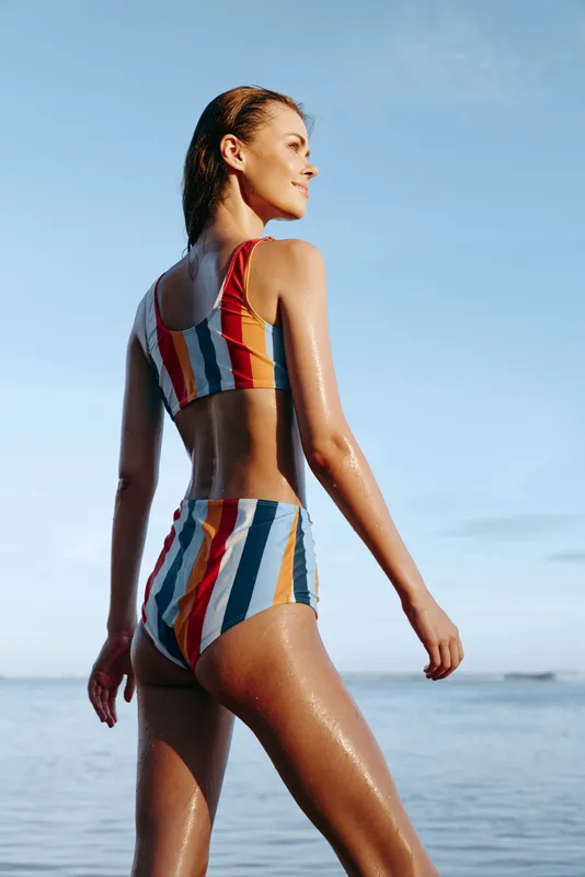 Young woman in a colorful striped swimsuit stands confidently by the calm sea under a clear blue sky, enjoying a sunny day at the beach.