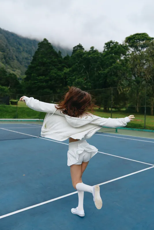 Young woman dancing joyfully on a tennis court, wearing a white outfit and socks, surrounded by green trees and mountains under a cloudy sky