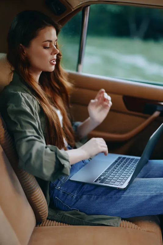 Young woman working on a laptop in a car, wearing casual attire and showing concentration, with a natural outdoor background in soft tones.