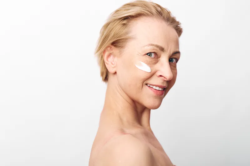 Smiling mature woman with short blonde hair applies white face cream on cheek. Close-up on white background. Concept of skincare, aging skin, beauty routine, and self-care for women.