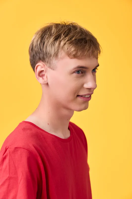 Boy, teen with short blond hair in a red shirt, side profile against a bright yellow background, casual pose, clean lighting, simple studio scene