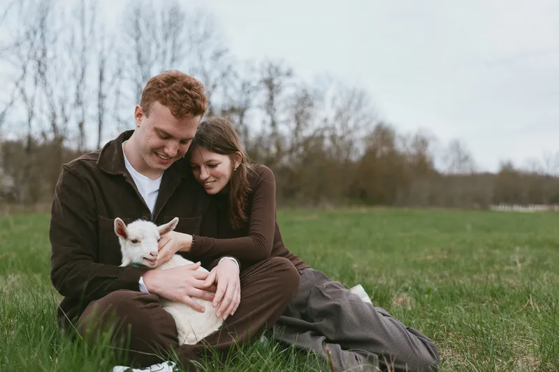 A couple sits on a grassy field, gently embracing a friendly goat kid in a bright outdoor setting. This lifestyle moment shows affection, warmth, and rural companionship.