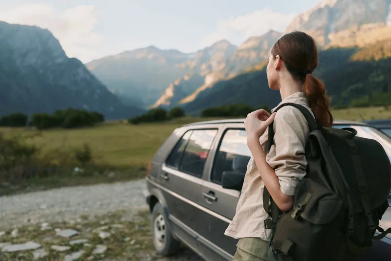 A woman with a backpack stands beside a parked car, gazing at rugged mountains and an open valley. A travel and outdoor lifestyle concept highlighting exploration and adventure.