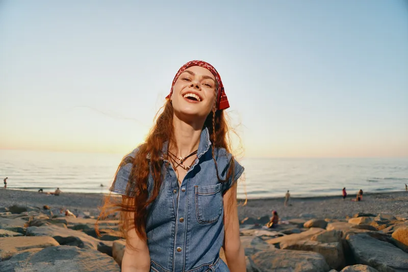 lifestyle woman wearing modern boho-western denim street style with red bandana, smiling joyfully outdoors at sunset by the beach in warm film color tones