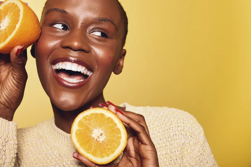 Happy African woman with dark skin holding two orange halves near her face smiling on yellow background. Concept for healthy lifestyle, fruit, vitamin c and natural beauty.
