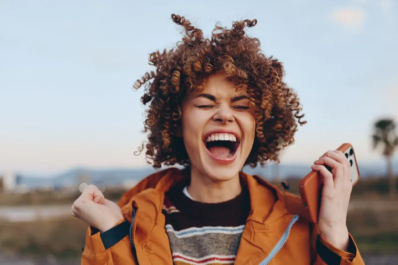 Happy cheerful laughing woman with curly hair in orange jacket, holding smartphone near face outdoors, sunny field landscape background, lifestyle communication, candid travel moment