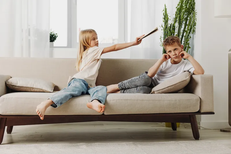 A playful girl in casual clothes hits a boy covering ears on a beige sofa in bright living room. Concept for sibling rivalry, childhood conflict, family dynamics, and home environment.