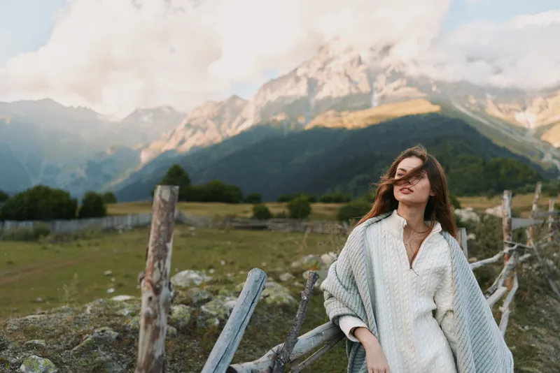 Cheerful woman outdoors wearing cozy cardigan near a rustic fence, mountain backdrop, soft sunlight, relaxed lifestyle moment, natural tones, lush landscape
