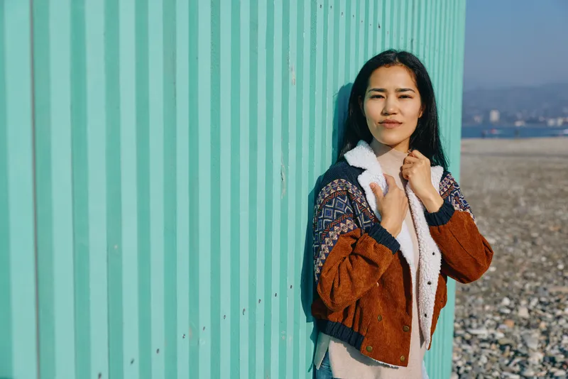 Woman leaning against a turquoise wall by a pebble beach, wearing a cozy shearling jacket, sunlight warming the scene, candid street style with a calm mood and subtle breeze