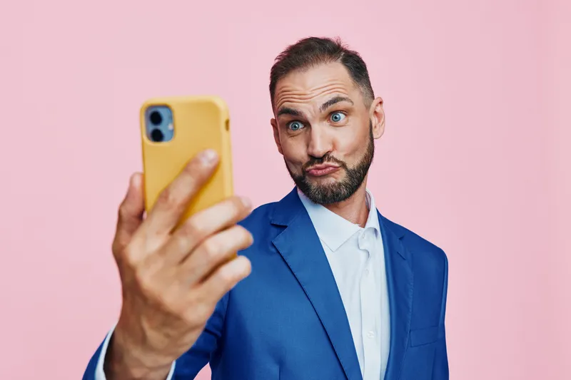 A professional man stands against a solid pink backdrop, an isolated color background. He poses for a selfie with a playful yet thoughtful expression, signaling confidence and curiosity.