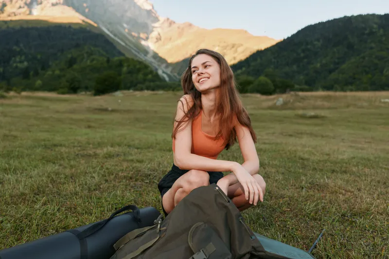 A cheerful outdoor traveler sits on a grassy meadow in a mountain landscape, smiling and relaxed next to a backpack and gear, enjoying a peaceful moment in nature.
