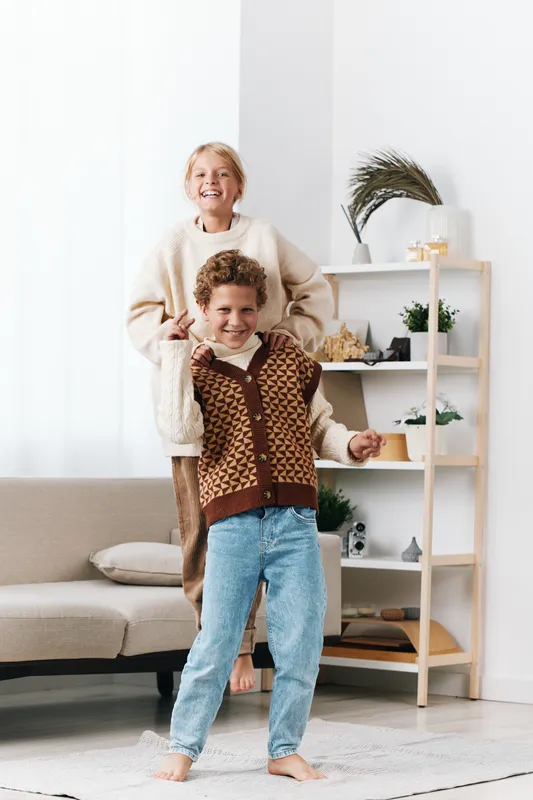 Two joyful children dance indoors in a bright, cozy living room, wearing warm sweaters. Soft natural light highlights their carefree, playful energy and joyful bond.