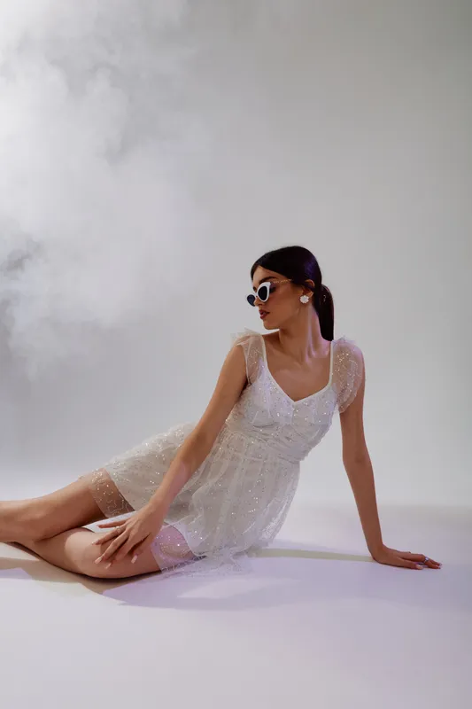Young woman in a white lace dress and sunglasses poses sitting on the floor against a smoky background. Concept of beauty, fashion, elegance, and modern style.