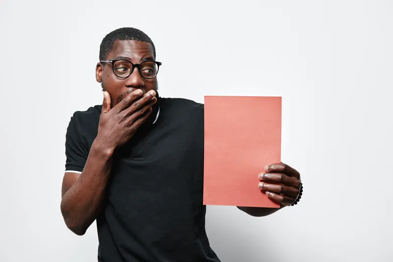 African American man with dark skin and glasses holding red paper with surprised expression on white background. Casual clothing and emotional reaction for concept or message.