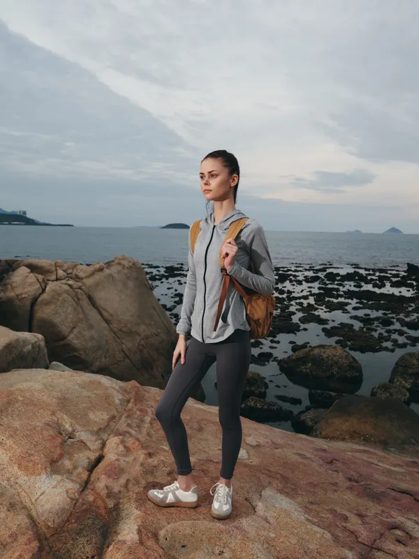 Young woman in activewear poses on rocky shore by the sea, showcasing a sporty lifestyle in nature, wearing a gray zip up jacket, gray leggings, and sneakers