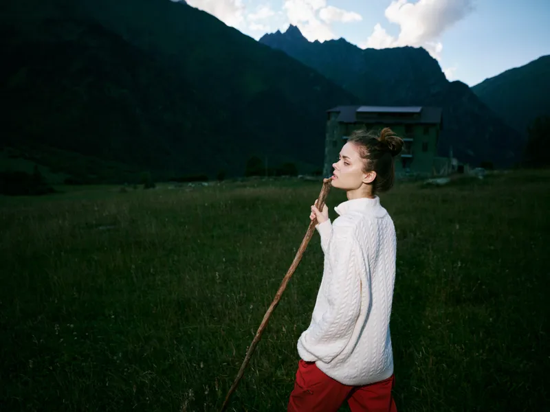 Woman walking through a grassy field with a long stick, mountains in the background, soft light, rural setting, distant valley, outdoor adventure and nature exploration scene