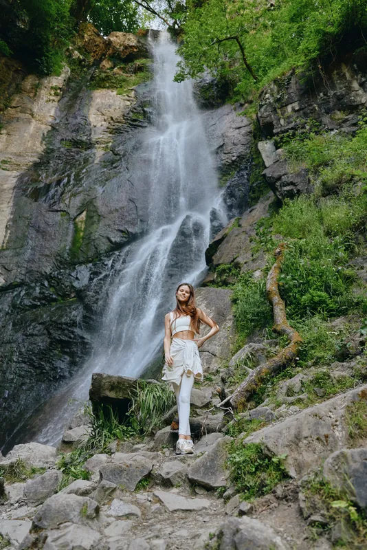 woman standing near waterfall in hiking outfit surrounded by lush green forest and rocky cliffs, serene nature scene, outdoor adventure